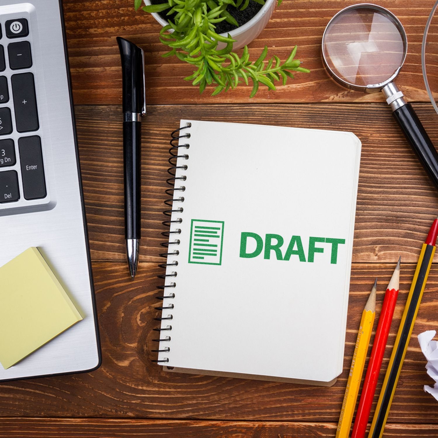 Self Inking Draft with Letter Stamp on a spiral notebook, surrounded by a laptop, pen, sticky notes, magnifying glass, and pencils on a wooden desk.