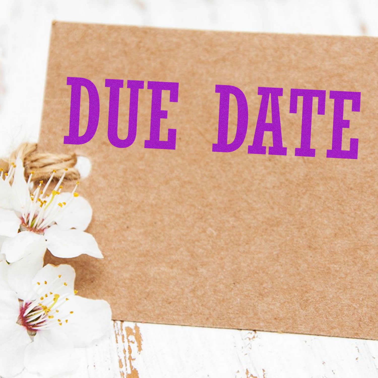 A brown card stamped with DUE DATE in purple ink using the Due Date Rubber Stamp, adorned with white flowers on a rustic white surface.