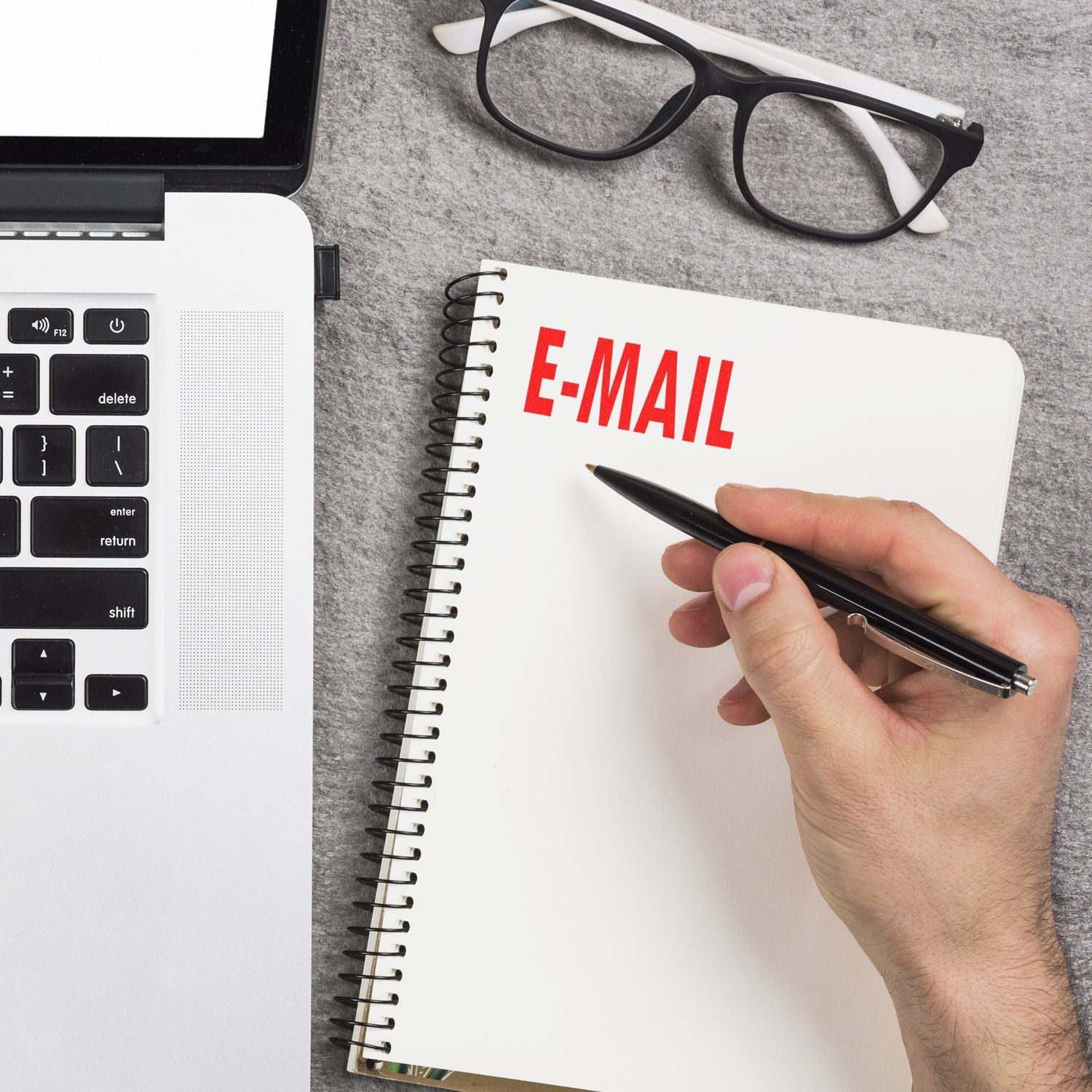 Hand holding a pen next to a notebook stamped with E-MAIL using the E Mail Rubber Stamp, placed beside a laptop and glasses on a gray surface.