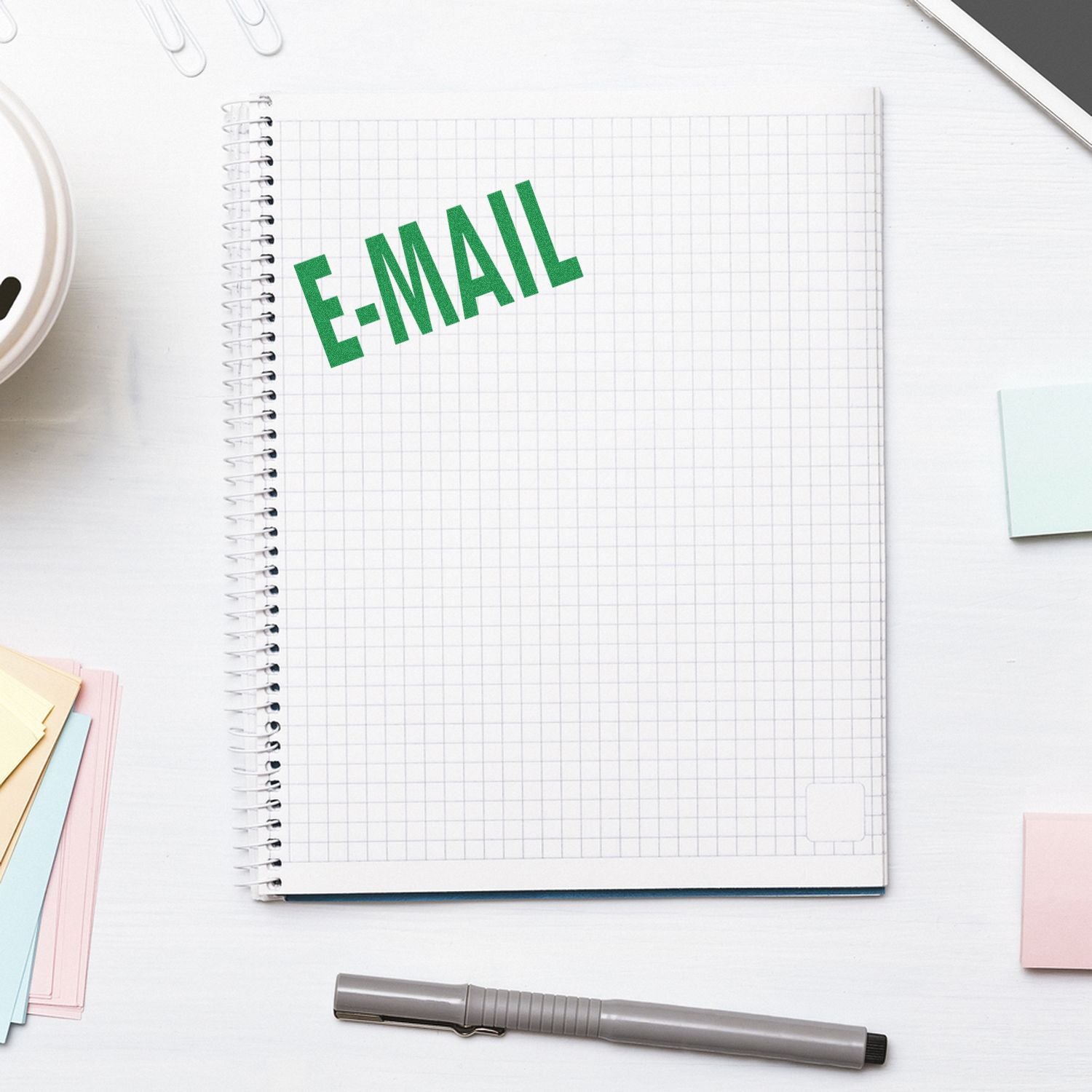 Large E Mail Rubber Stamp in green ink on a spiral notebook, surrounded by a pen, sticky notes, and a smartphone on a white desk.