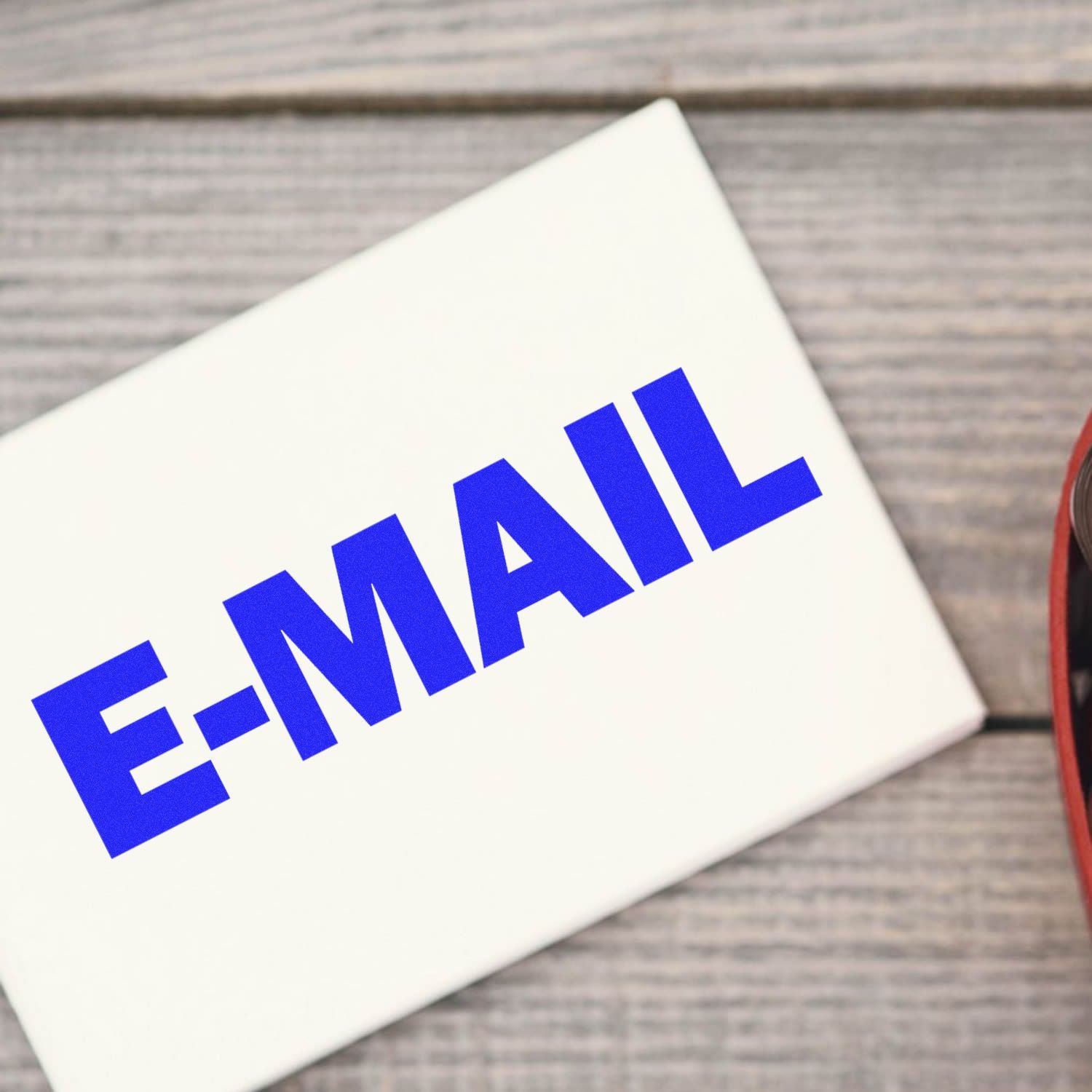 E-Mail Rubber Stamp in use, showing a bold blue E-MAIL imprint on a white surface, placed on a wooden background.