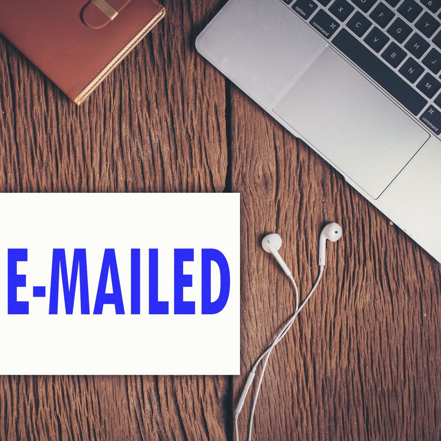 Slim Pre-Inked E-Mailed Stamp in blue on a white paper, next to a laptop, earphones, and a brown notebook on a wooden desk.