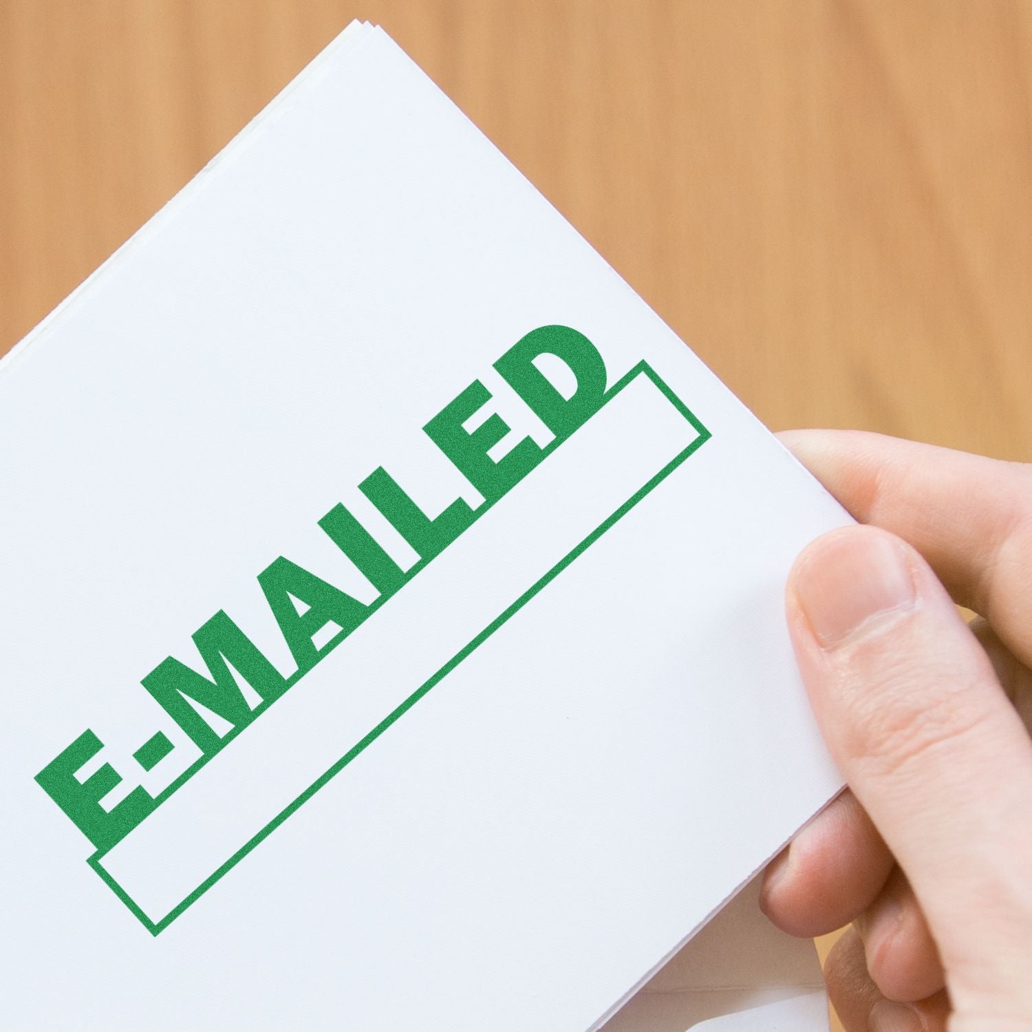 Hand holding a white paper stamped with E-MAILED in green using the E-mailed with Date Box Rubber Stamp, on a wooden background.