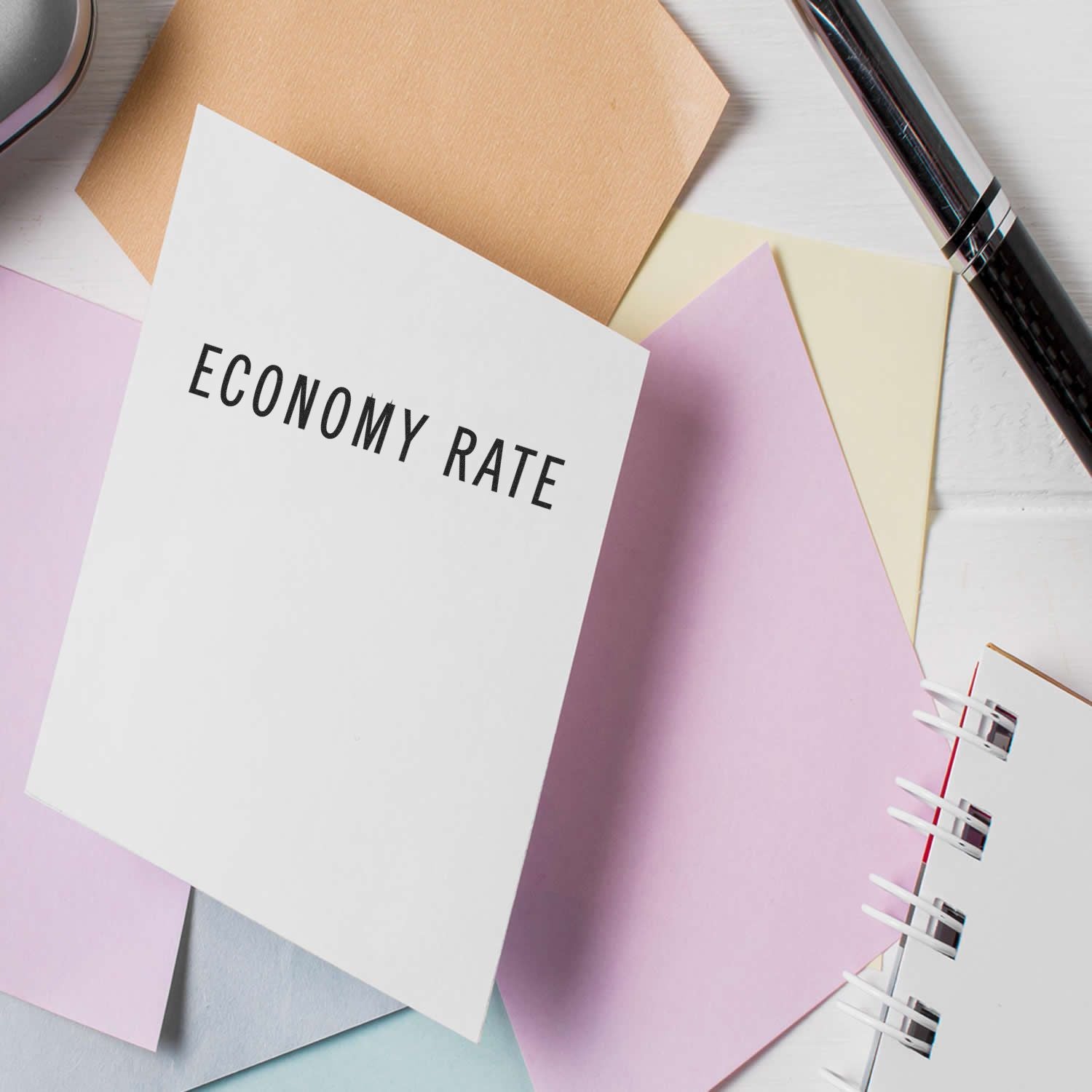 Economy Rate Rubber Stamp used on a white card, surrounded by pastel-colored papers, a pen, and a notebook on a white surface.