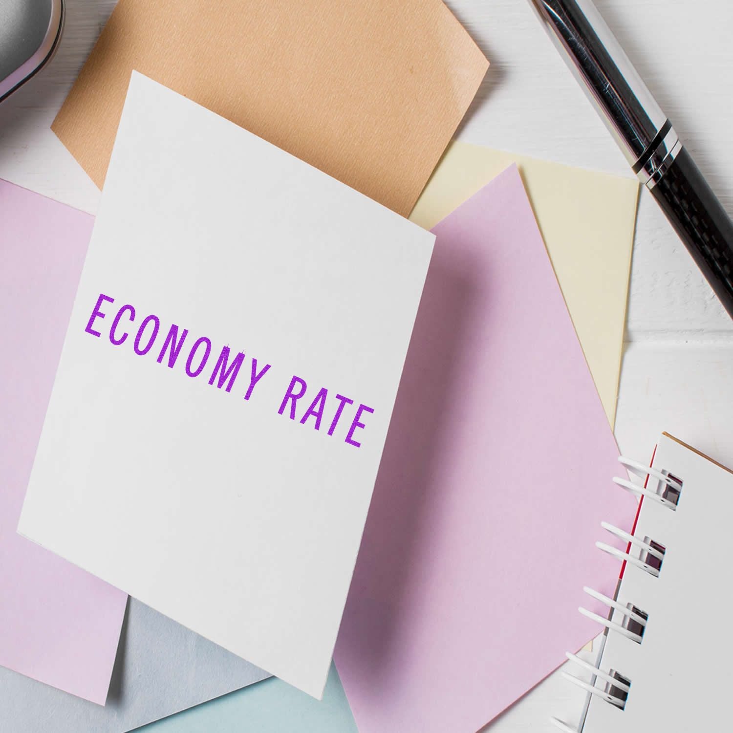 Economy Rate Rubber Stamp in use on a white card, surrounded by pastel-colored papers, a pen, and a notebook on a white desk.