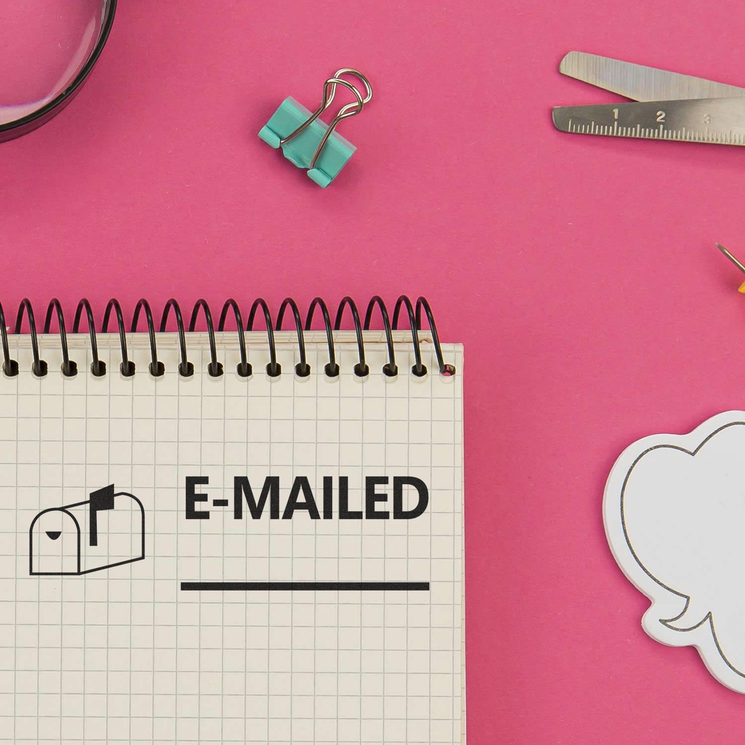 Emailed with Mailbox Rubber Stamp used on a spiral notebook page, surrounded by office supplies on a pink background.