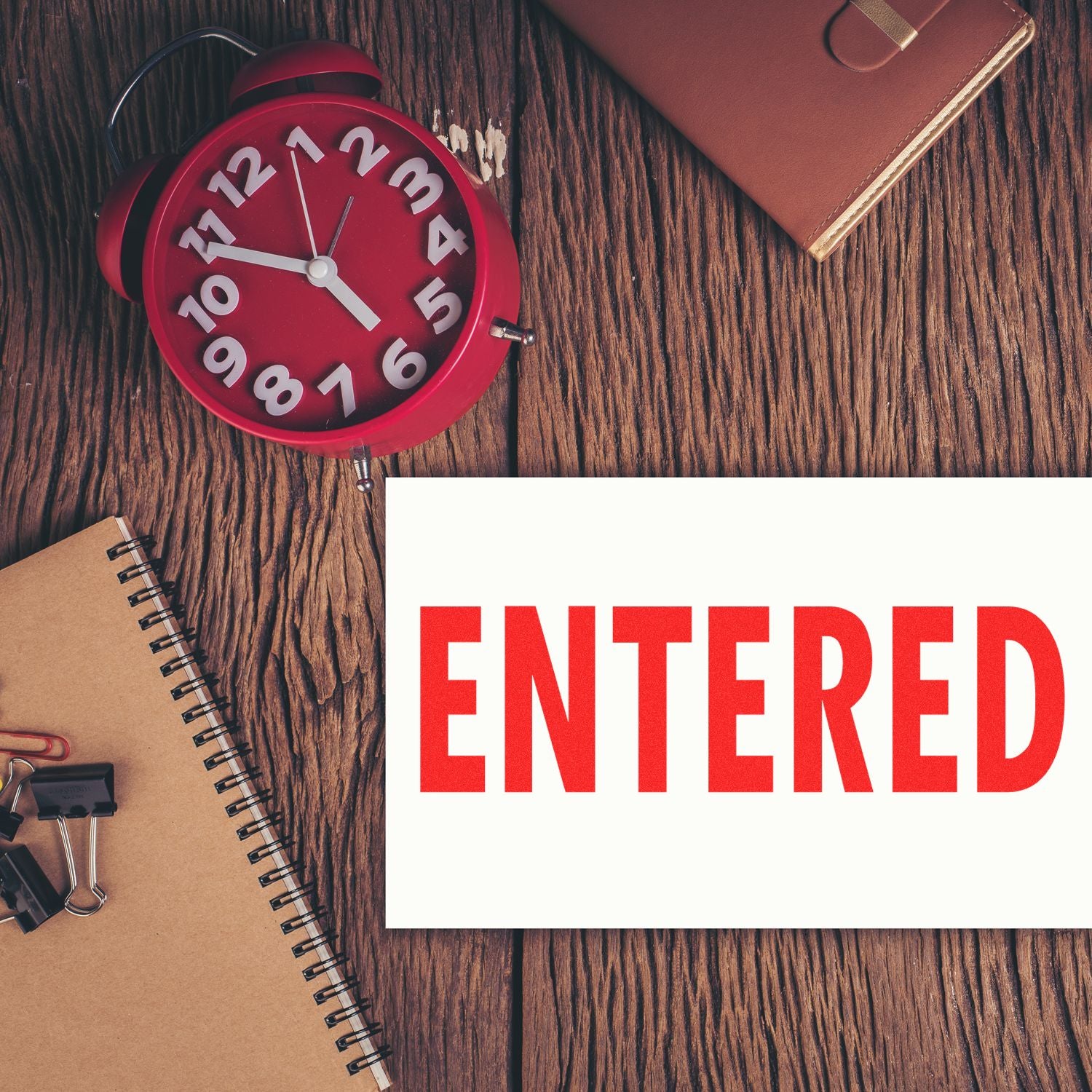 Large Entered Rubber Stamp in red ink on white paper, placed on a wooden desk with a red clock, notebook, and office supplies.