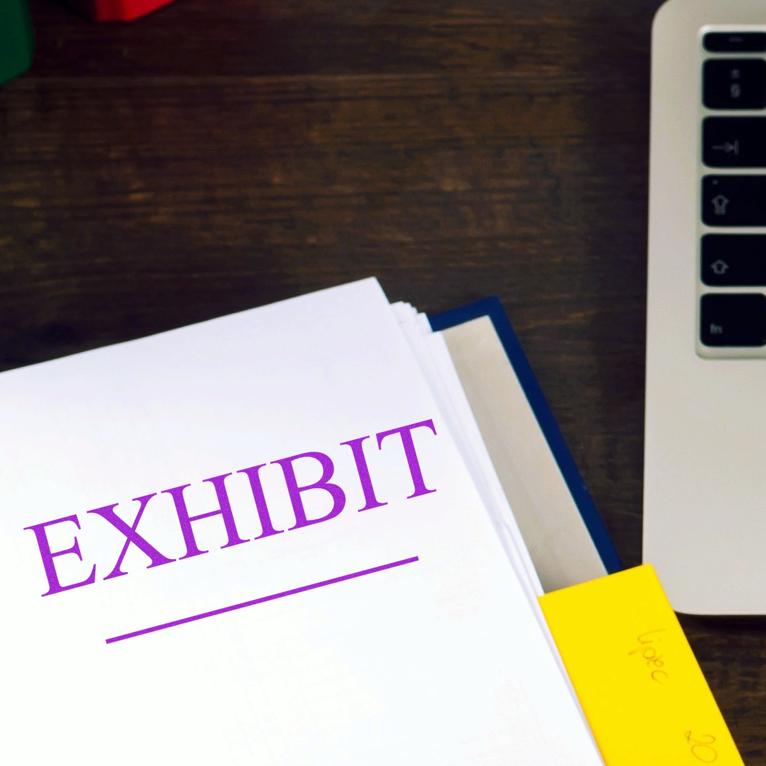 Exhibit rubber stamp marking on a document next to a laptop and sticky notes on a wooden desk.