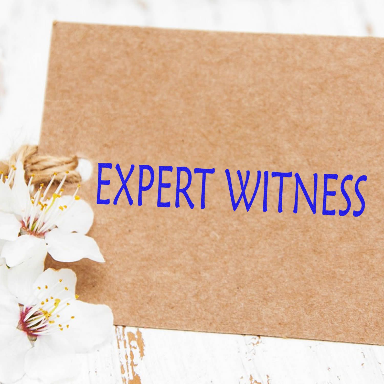 A Slim Pre-Inked Expert Witness Stamp on a brown card with white flowers on a rustic white wooden surface.