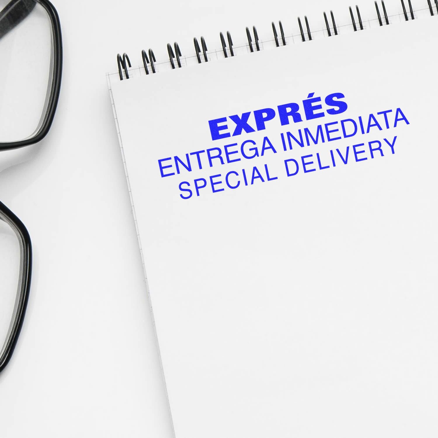 Self Inking Expres Entrega Inmedia Stamp in blue ink on a white notepad, next to black-framed glasses.