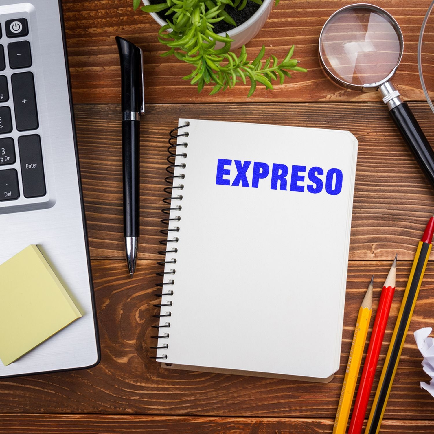 Expreso Rubber Stamp in use on a spiral notebook, placed on a wooden desk with a laptop, pen, sticky notes, magnifying glass, and pencils.