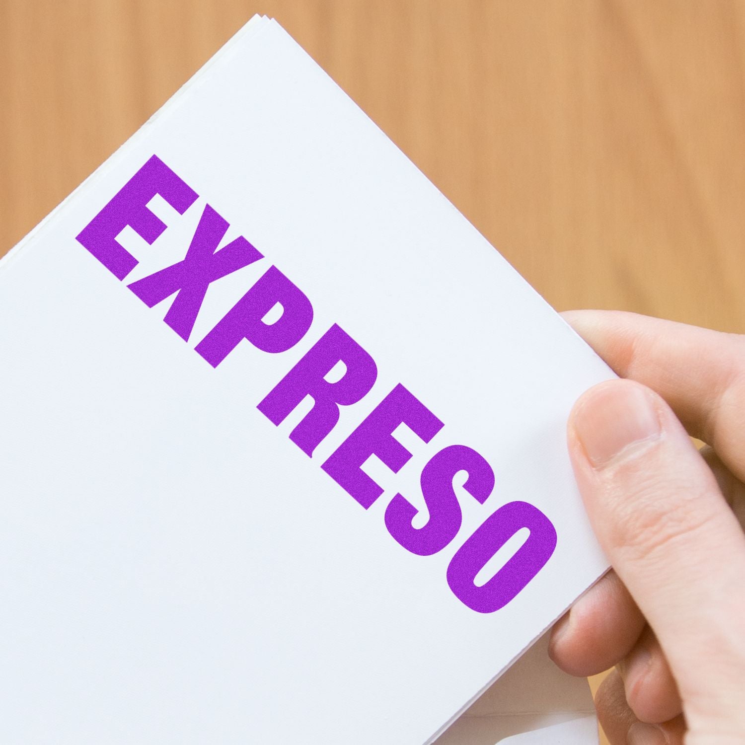 Hand holding a white card stamped with EXPRESO in purple ink using the Large Pre-Inked Expreso Stamp against a wooden background.