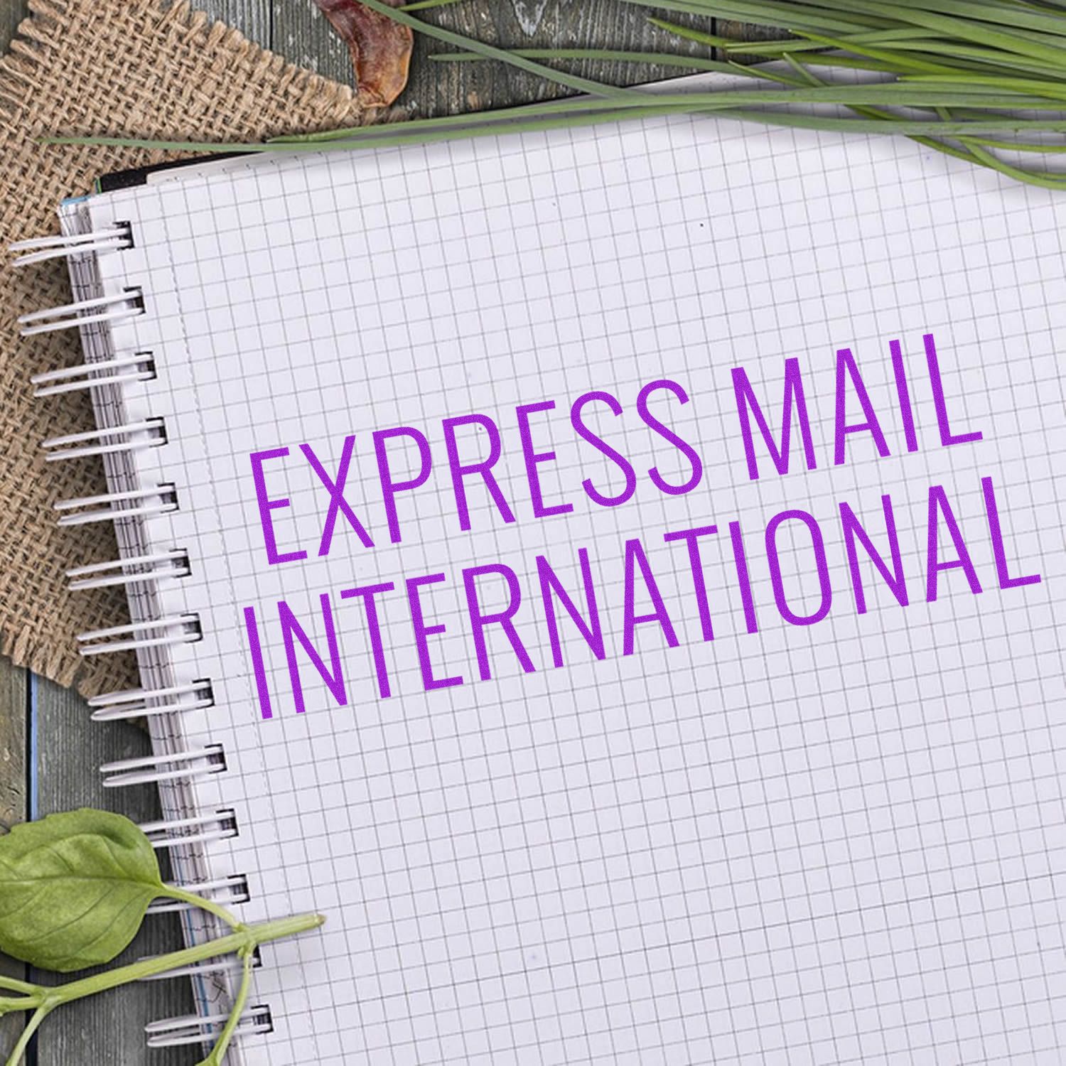 Large Pre-Inked Express Mail International Stamp on a spiral notebook with grid paper, surrounded by leaves and burlap fabric.