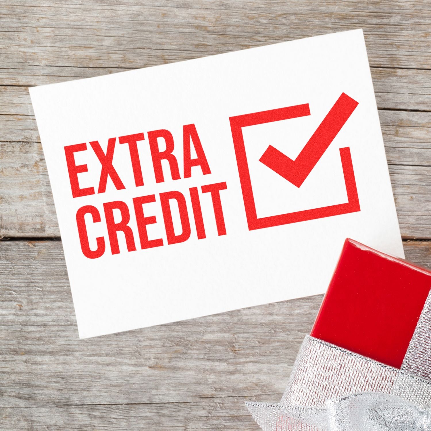 Extra Credit Rubber Stamp in use on a white card with a red checkmark, placed on a wooden surface next to a wrapped gift.