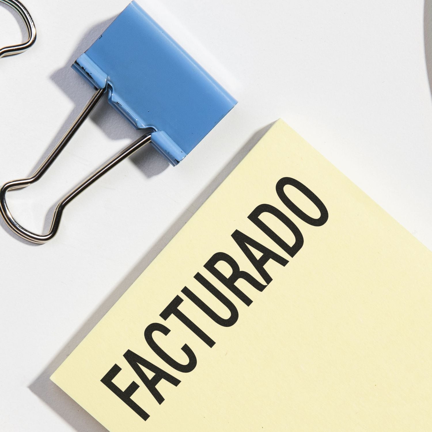 Facturado Rubber Stamp in use on a yellow sticky note, next to a blue binder clip on a white surface.