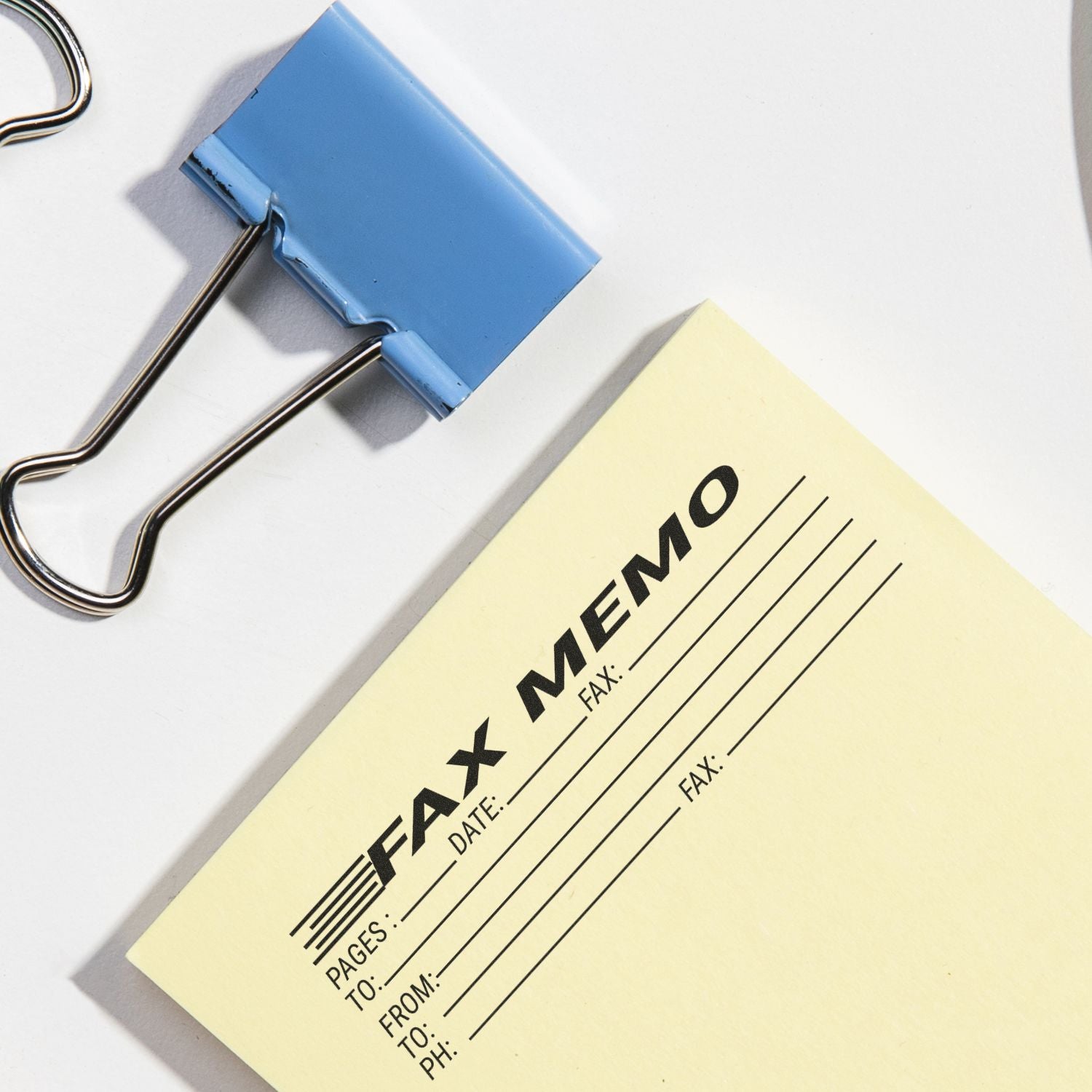 A blue binder clip and a yellow paper stamped with Fax Memo Rubber Stamp showing fields for pages, date, to, from, and fax number.