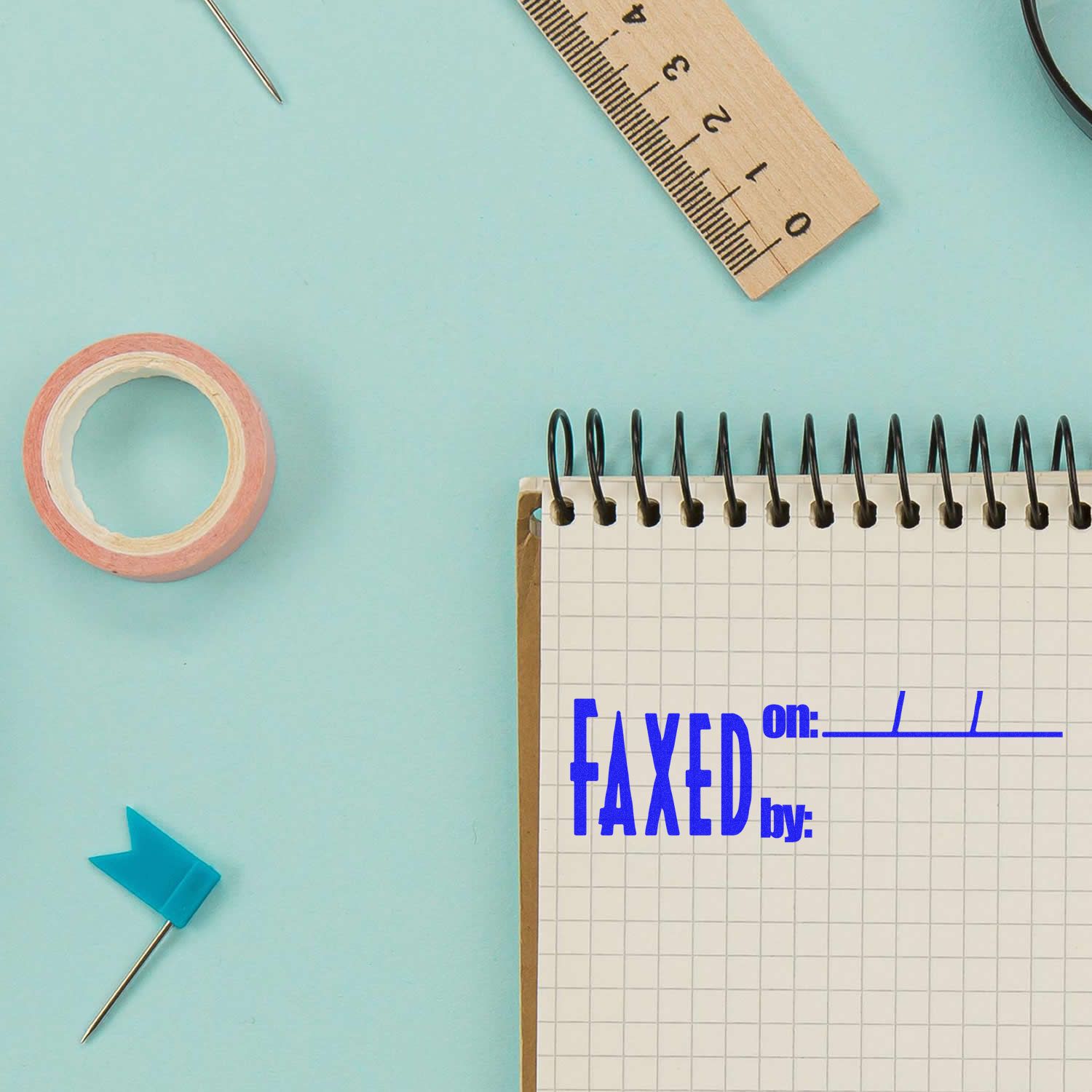 A Large Faxed on Rubber Stamp is used on a spiral notebook with office supplies like a ruler, tape, and a pushpin on a light blue background.