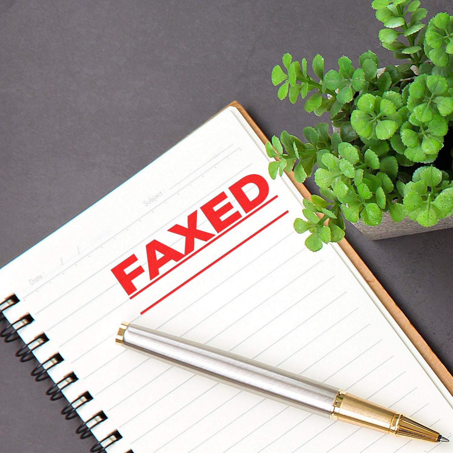 Notebook with FAXED stamped in red using the Faxed with Line Rubber Stamp, next to a silver pen and a small potted plant on a gray surface.