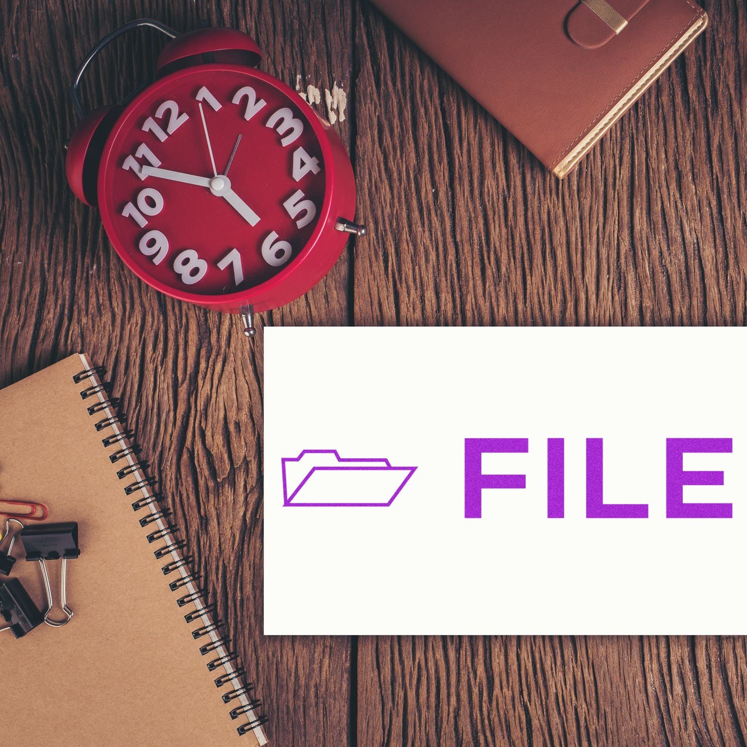 A red clock, notebook, and binder clips on a wooden desk with a card stamped with File with Folder Rubber Stamp showing a folder icon and the word FILE .