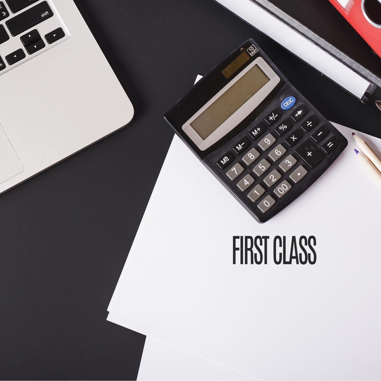 A Large First Class Rubber Stamp is used on a stack of white papers next to a calculator, laptop, and pencil on a black desk.