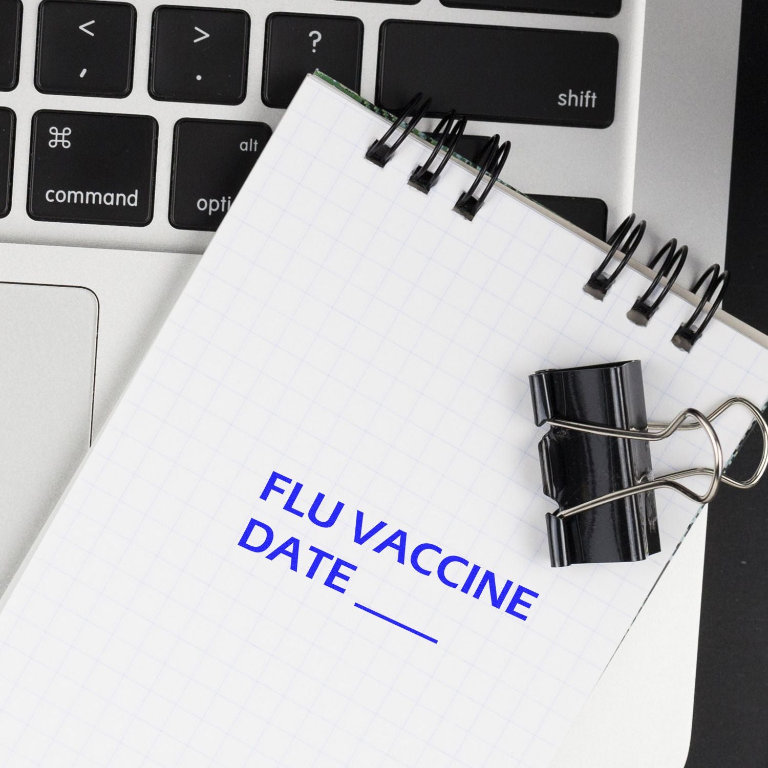 A Large Pre-Inked Flu Vaccine Date Stamp is shown on a notepad with a laptop keyboard in the background.