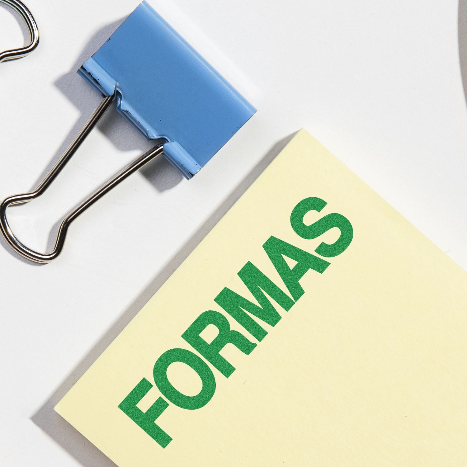 A blue binder clip and a yellow notepad stamped with "FORMAS" using the Formas Rubber Stamp are shown on a white surface.