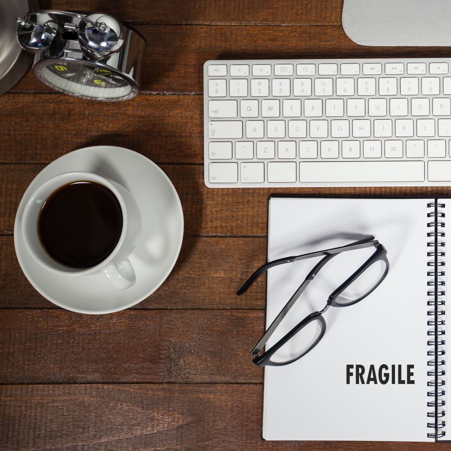 Desk with a keyboard, coffee cup, glasses, and a notebook stamped with 'Fragile Rubber Stamp' on a wooden surface.