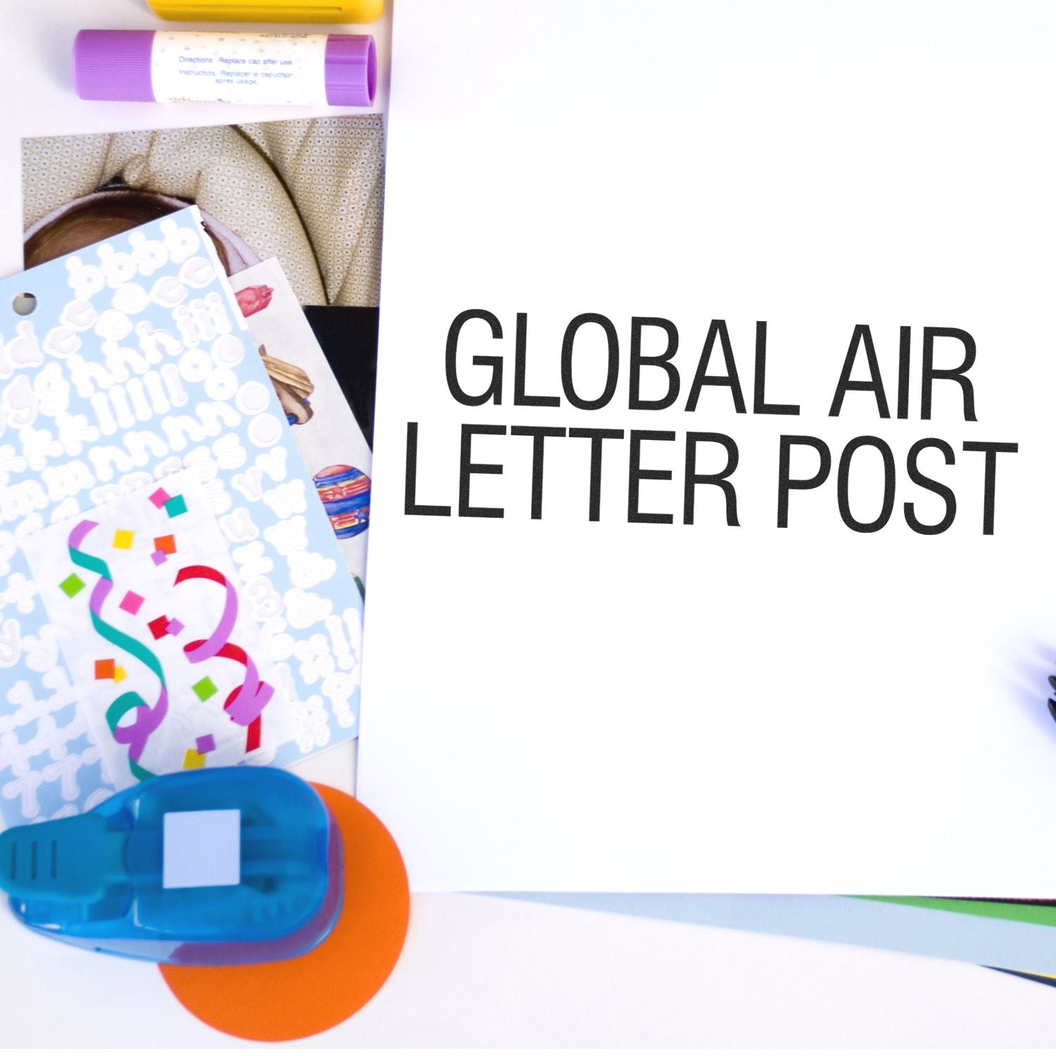 Desk with stationery items and a Large Global Air Letter Post Rubber Stamp on a white paper, surrounded by stickers, markers, and a hole punch.