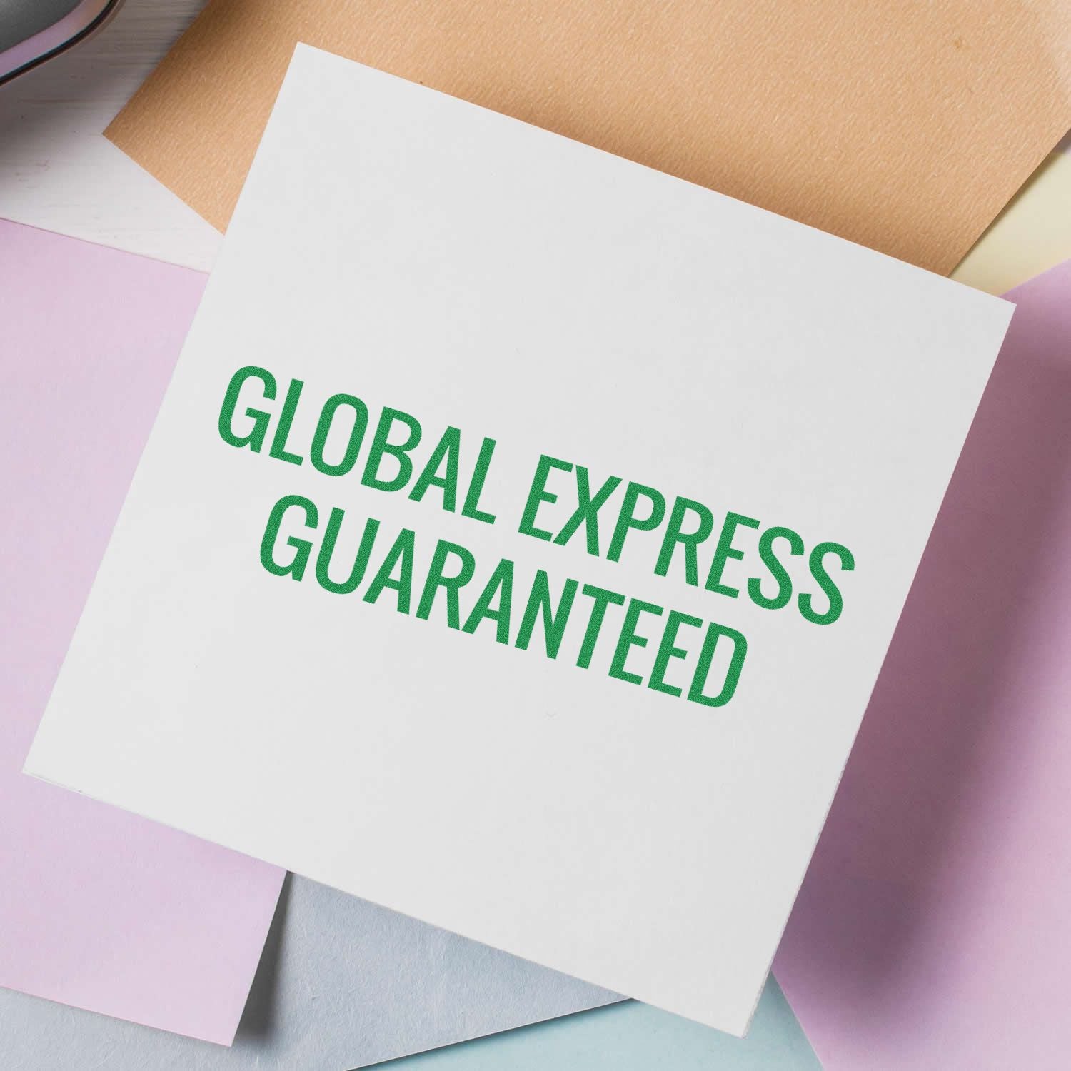 Global Express Guaranteed Rubber Stamp in use, stamping a white paper with green ink, surrounded by pastel-colored papers.