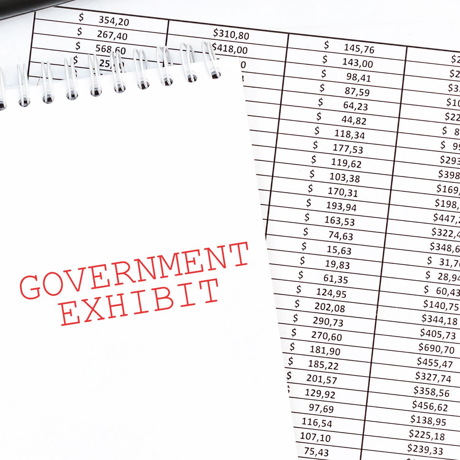 A Government Exhibit Rubber Stamp is used on a spiral notebook, placed on a table with a financial spreadsheet in the background.