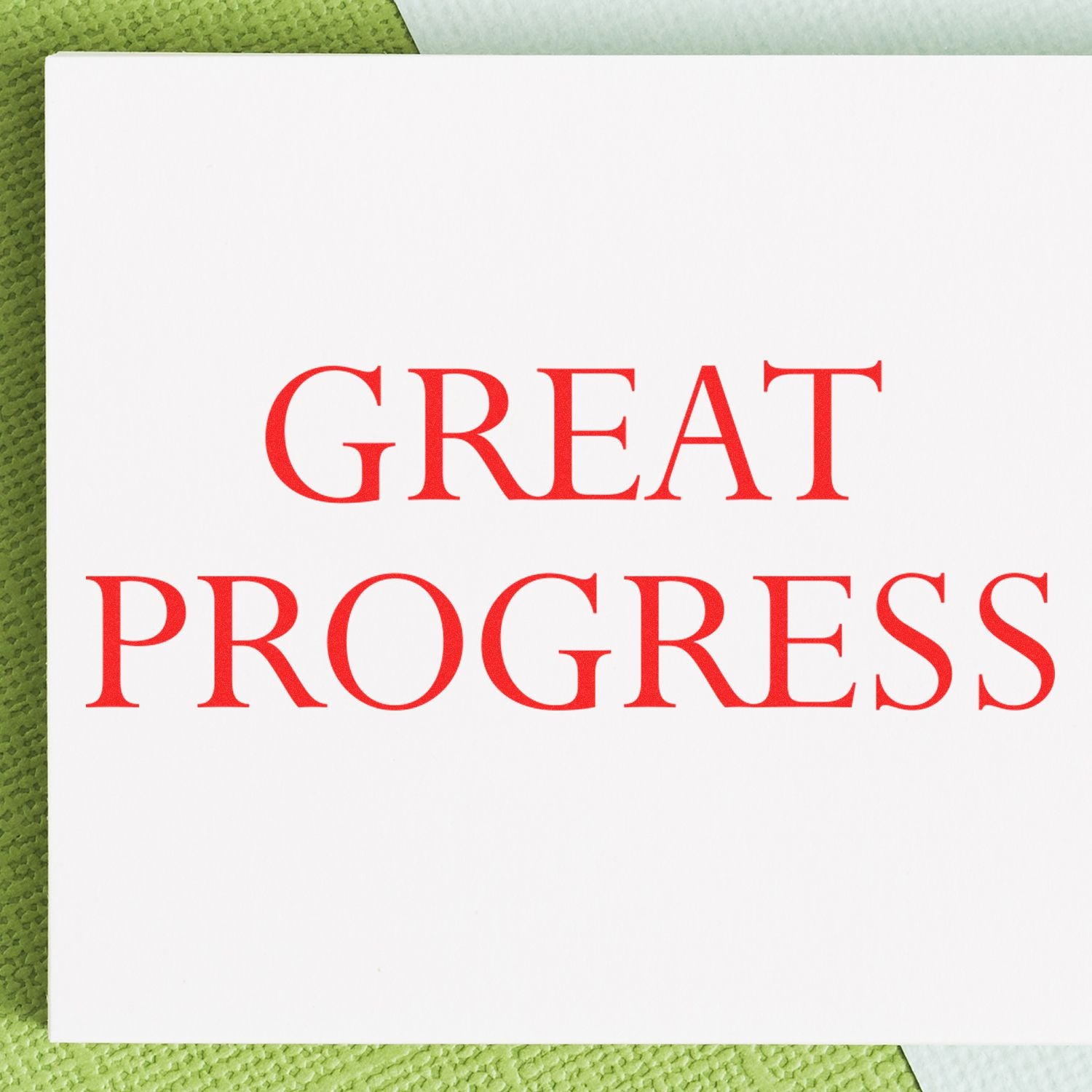 A white card stamped with GREAT PROGRESS in red ink using the Large Pre-Inked Great Progress Stamp, placed on a green textured surface.