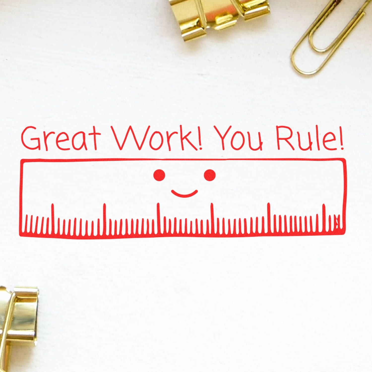 Great Work You Rule Rubber Stamp in red ink on white paper, featuring a smiling ruler design. Gold binder clips and paper clips are nearby.