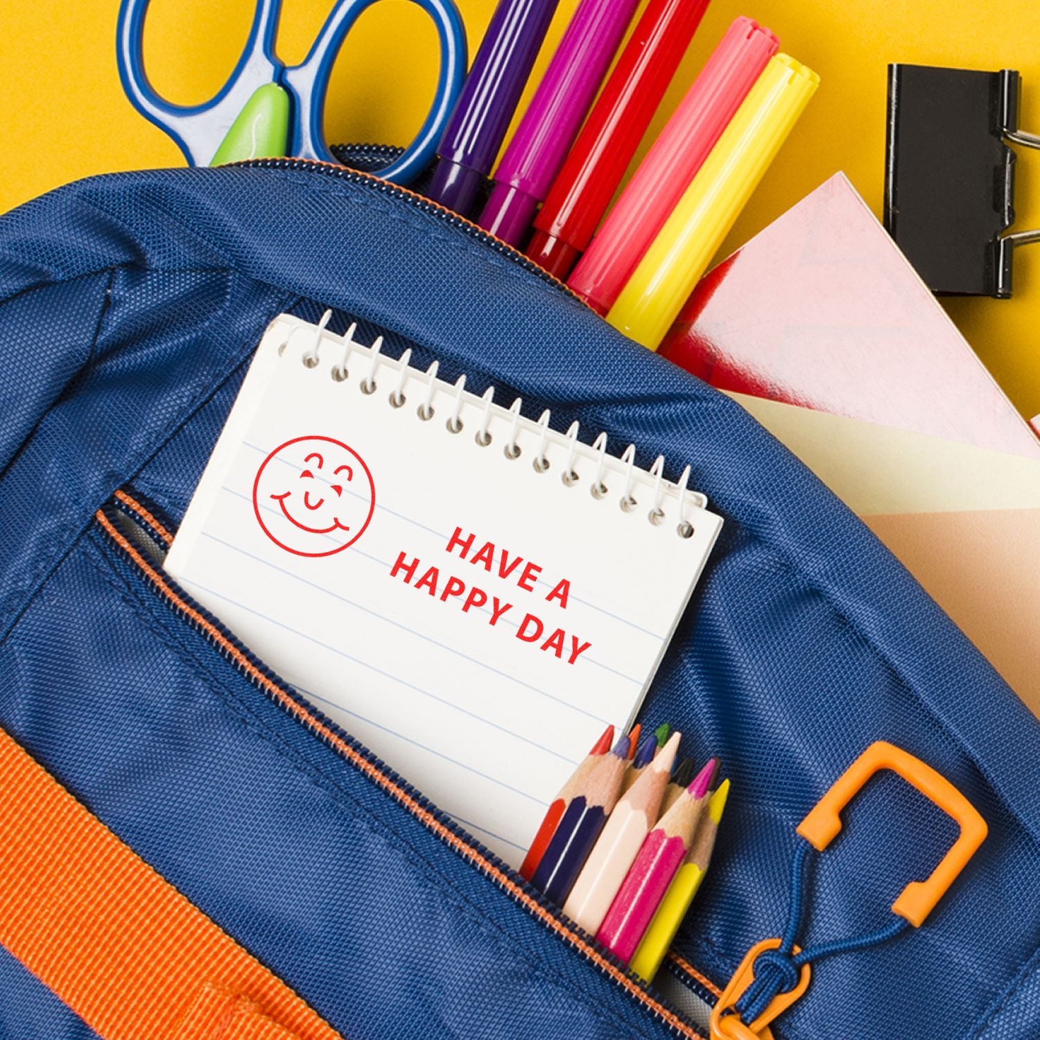 Slim Pre-Inked Have a Happy Day Stamp on a notepad inside a blue backpack with colorful pens, pencils, and scissors.