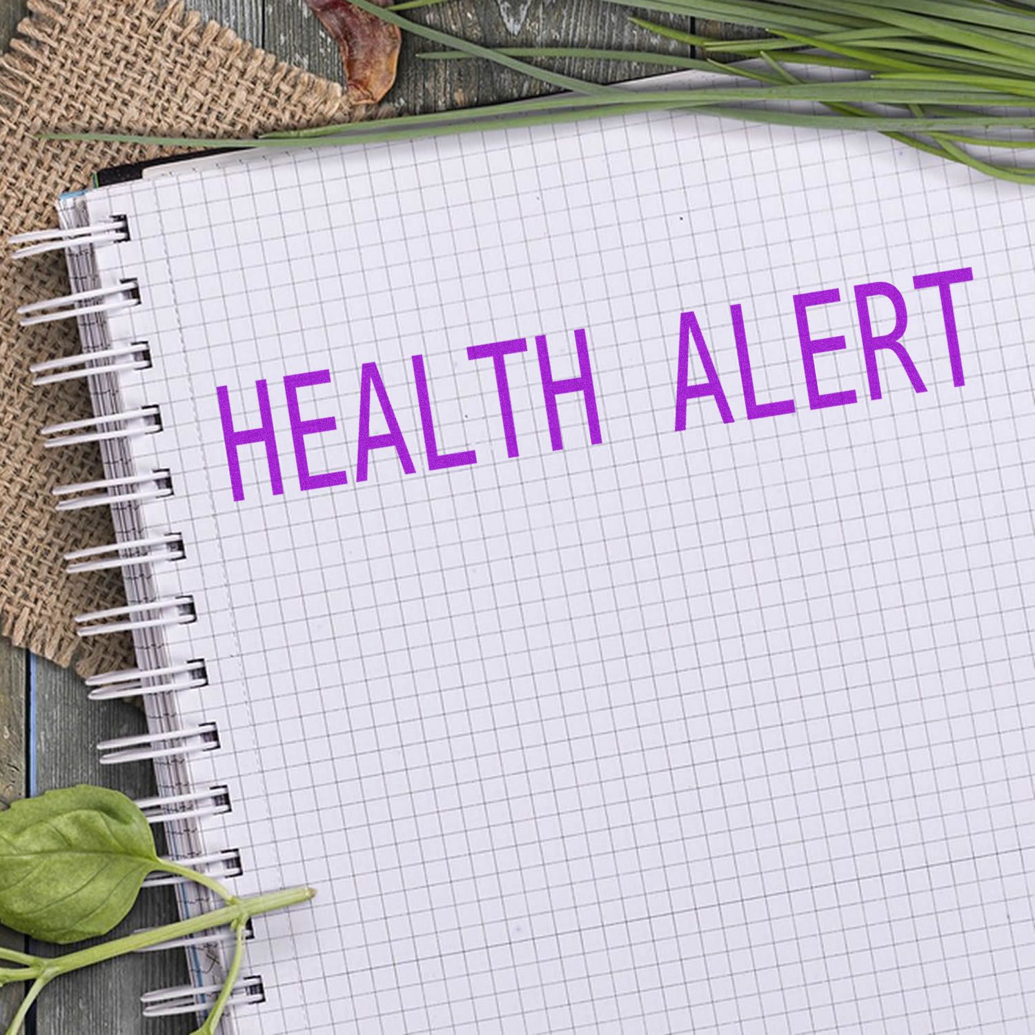 Notebook with HEALTH ALERT stamped in purple using the Health Alert Rubber Stamp, surrounded by leaves and a burlap cloth on a wooden surface.