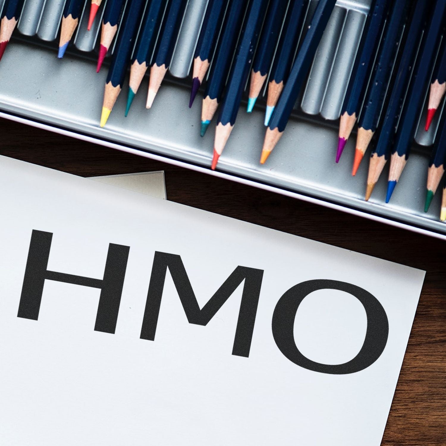 HMO Medical Rubber Stamp in use, with colorful pencils in the background. The stamp imprint reads HMO on a white sheet of paper.