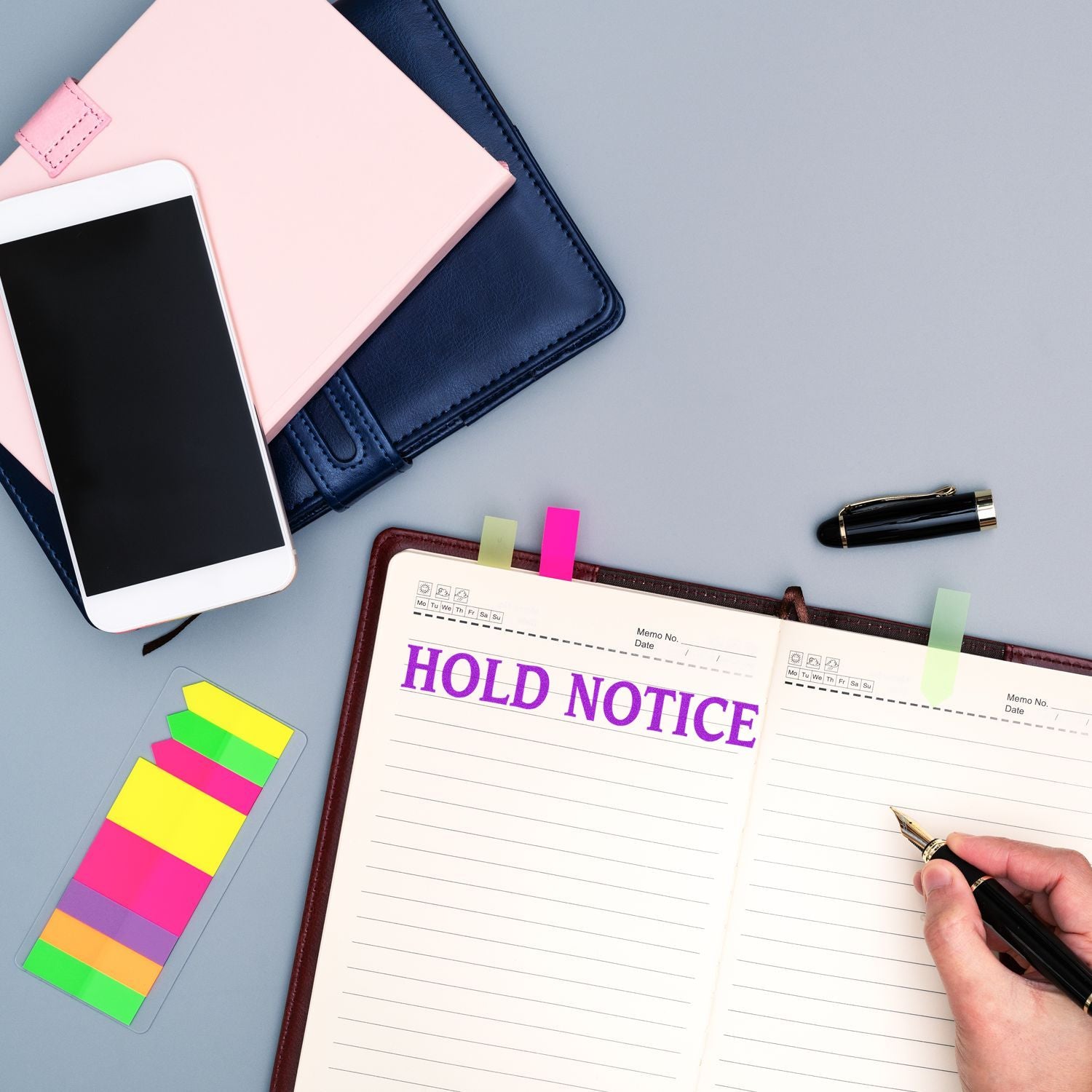 Person using a Large Hold Notice Rubber Stamp on a notebook, surrounded by a smartphone, colorful sticky notes, and a pen.