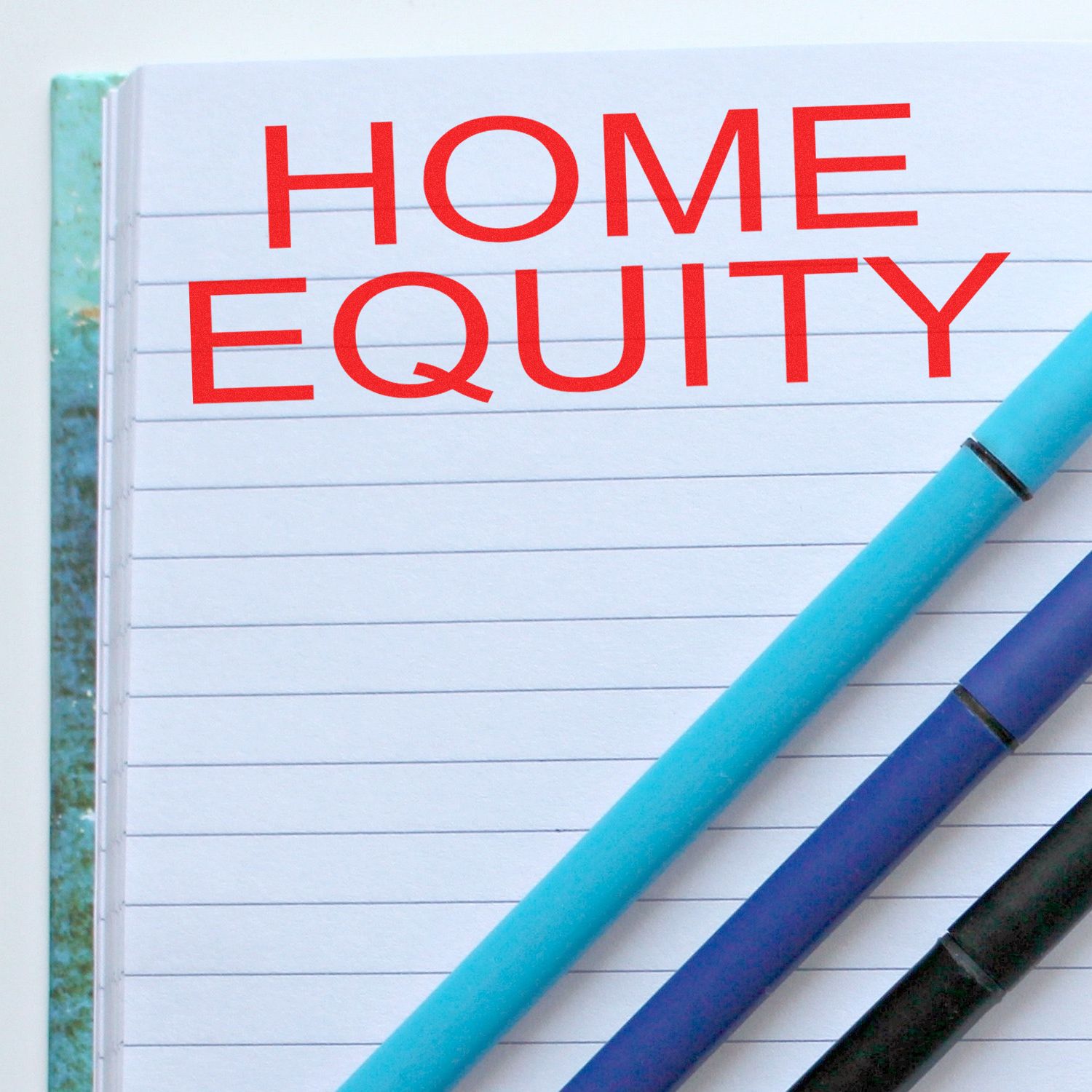 Home Equity Rubber Stamp in red ink on a lined notebook page, with three pens (blue, black, and turquoise) placed diagonally below the text.