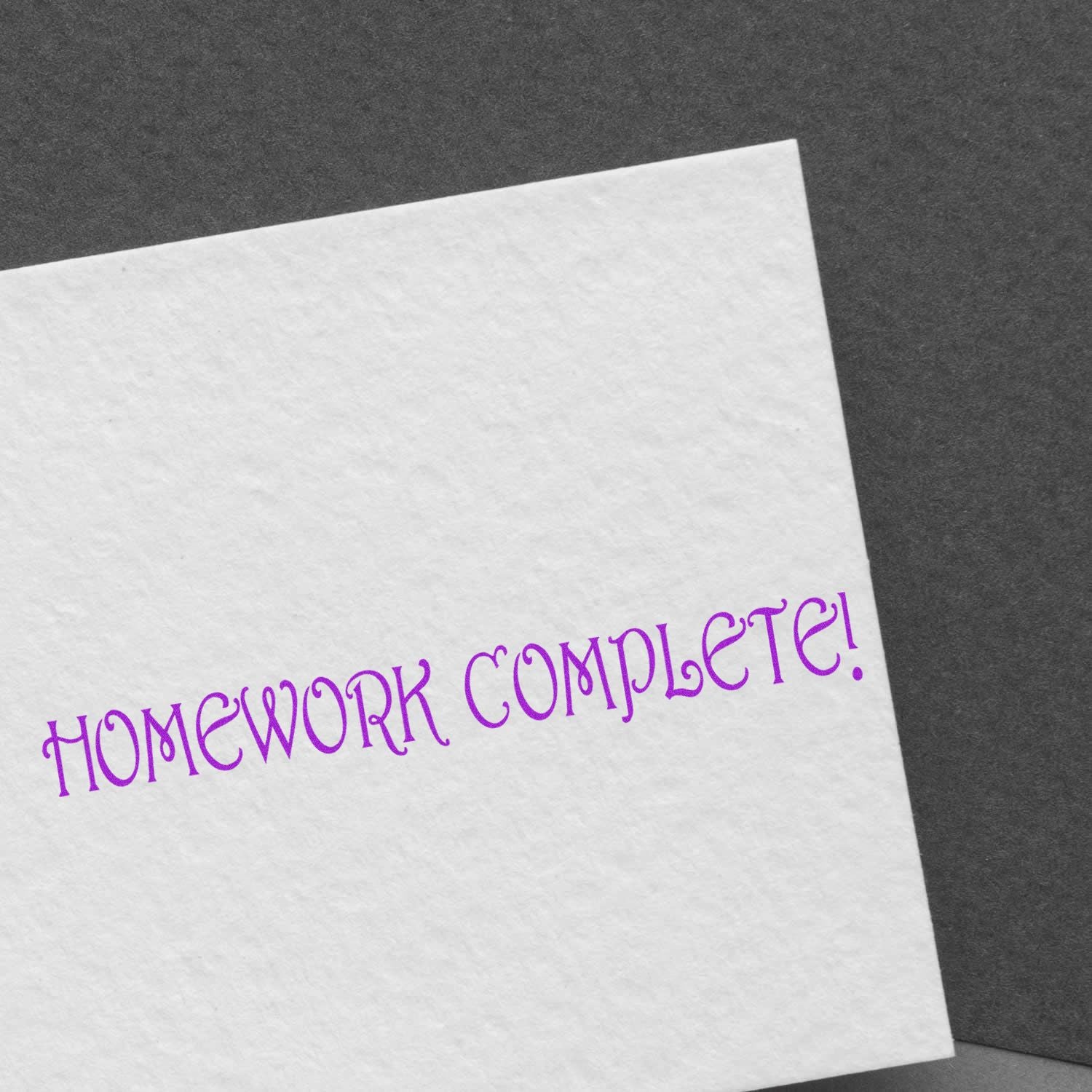 White paper stamped with HOMEWORK COMPLETE! in purple ink using the Large Homework Complete Rubber Stamp, on a dark background.