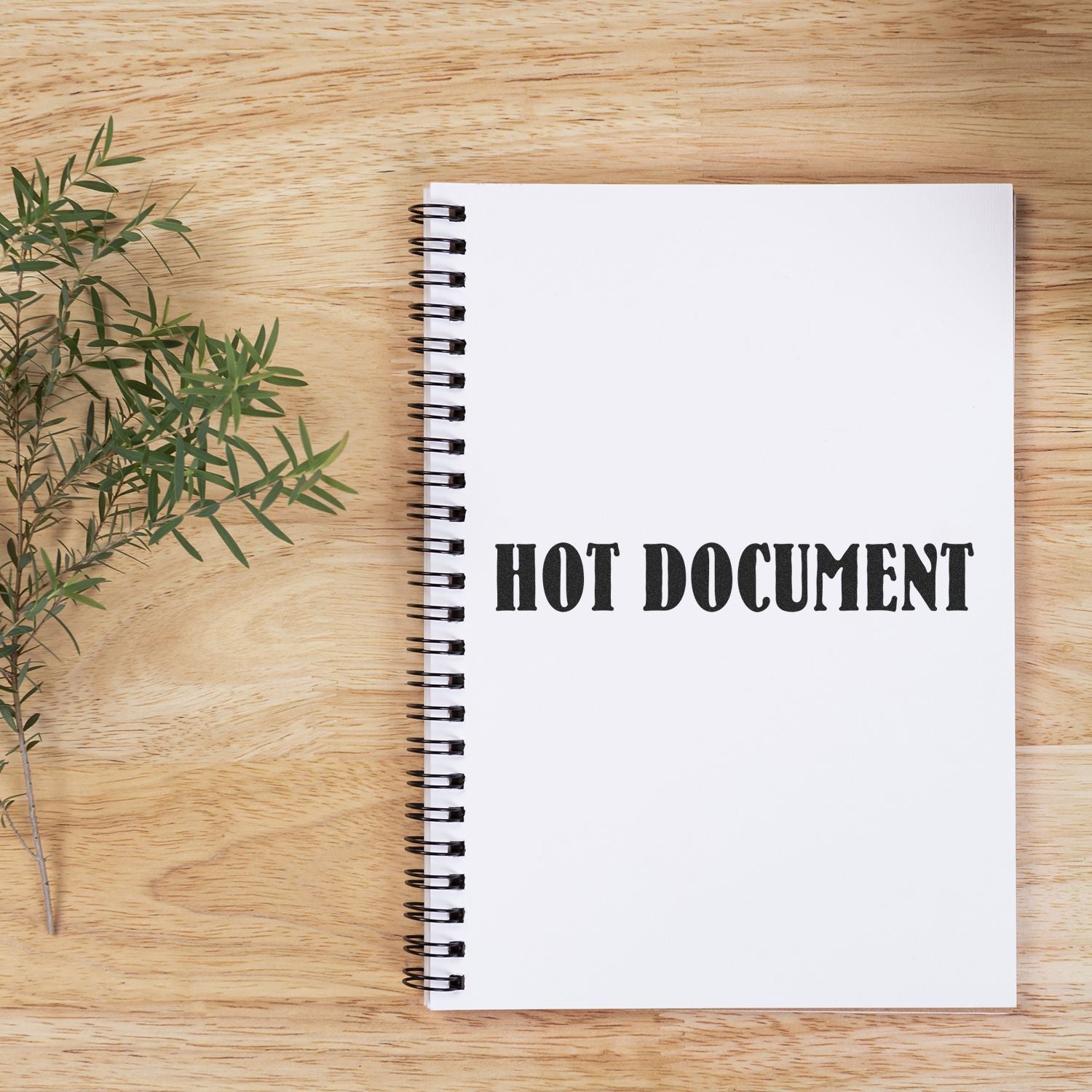 A spiral notebook on a wooden table stamped with HOT DOCUMENT using the Hot Document Rubber Stamp, next to a sprig of greenery.