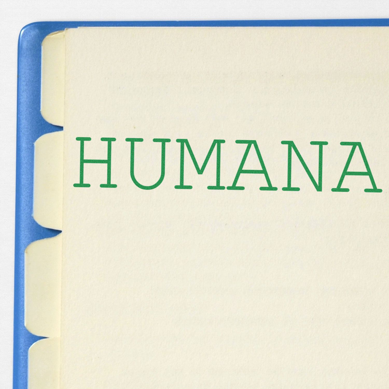 Humana Rubber Stamp in use, displaying the word HUMANA in green ink on a cream-colored paper with tabbed dividers in a blue binder.