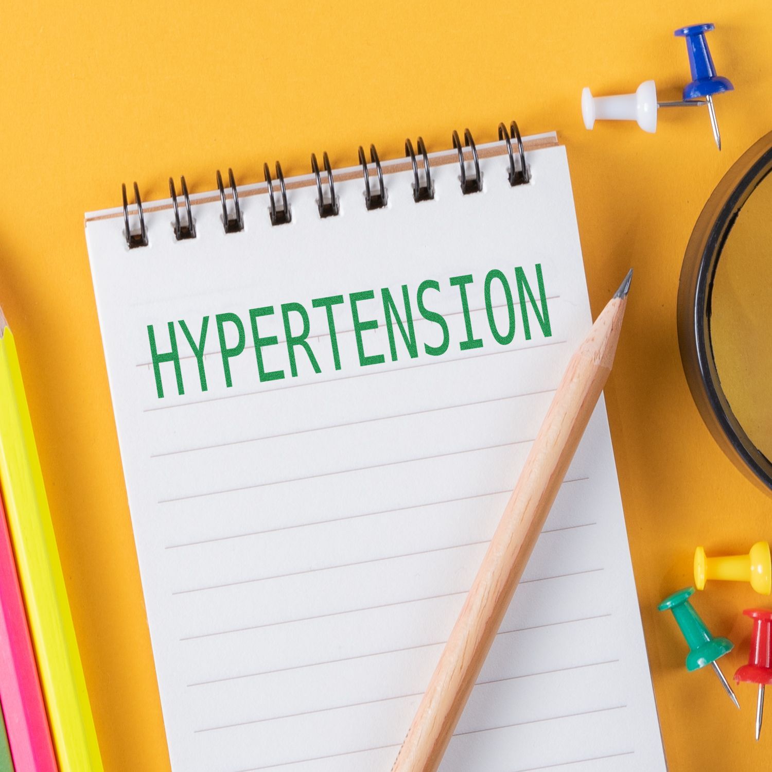 Notepad with HYPERTENSION stamped in green using the Hypertension Rubber Stamp, surrounded by a pencil, colorful pins, and a magnifying glass.