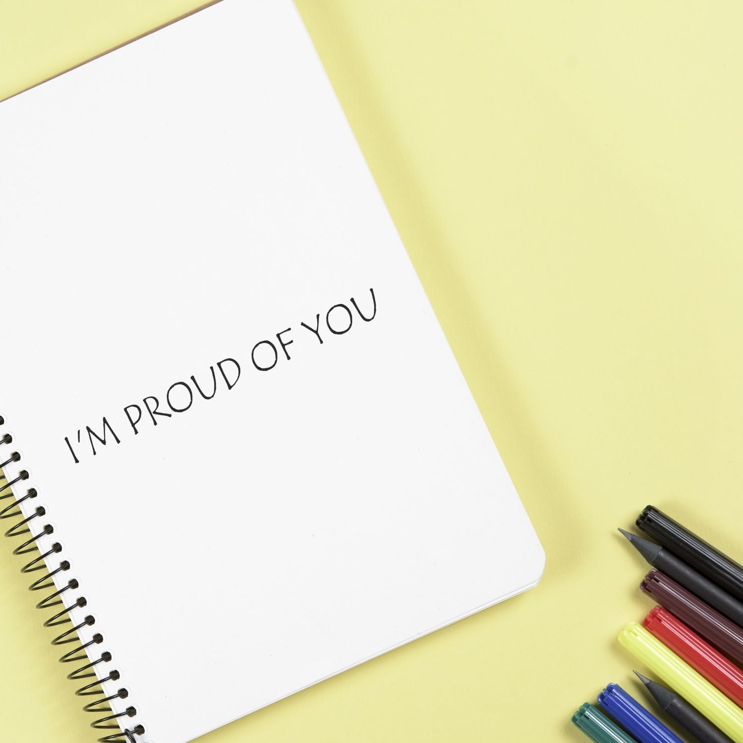 A notebook with I'M PROUD OF YOU stamped using the I'm Proud Of You Rubber Stamp, surrounded by colorful pens on a yellow background.