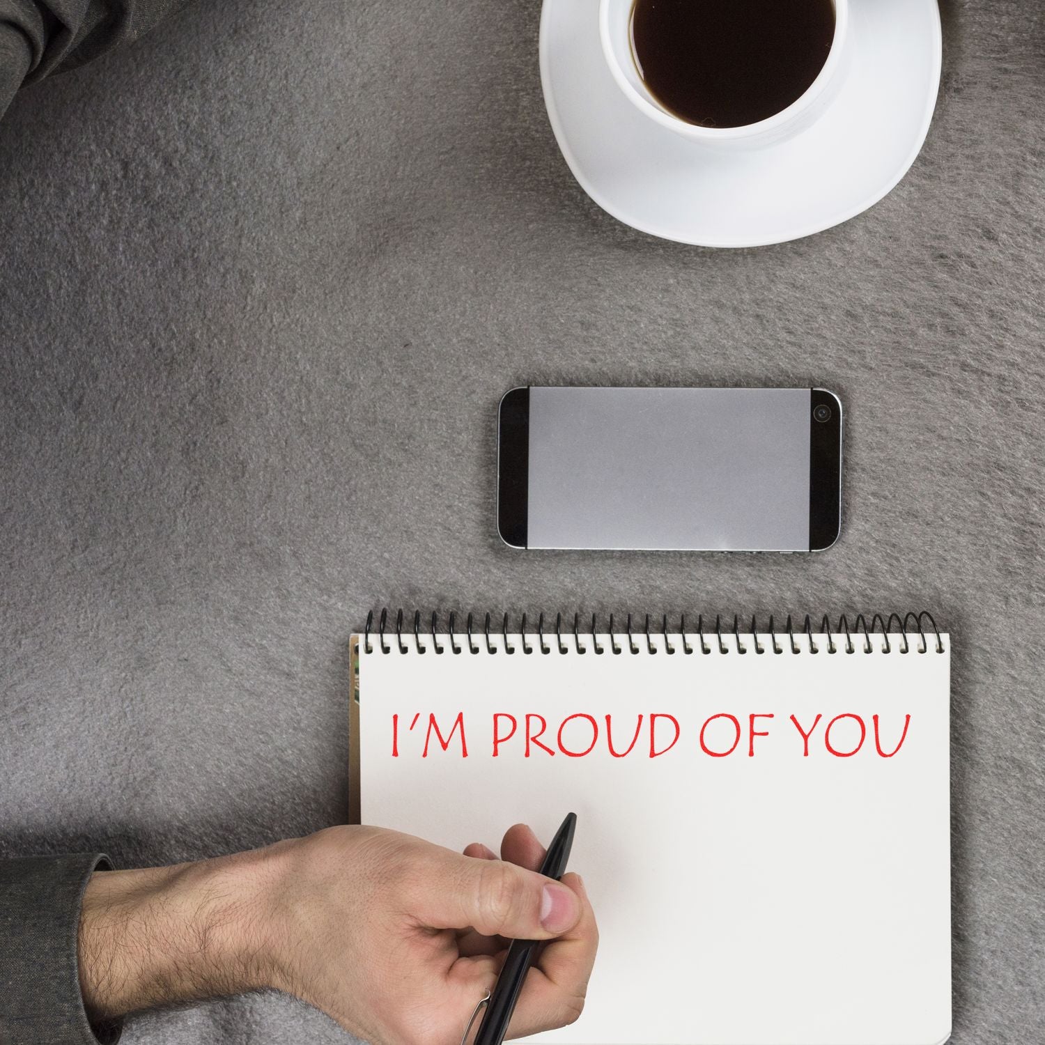 A hand holding a pen near a notebook stamped with I'M PROUD OF YOU in red ink, next to a phone and coffee cup. Product: Large Pre-Inked I'm Proud Of You Stamp.