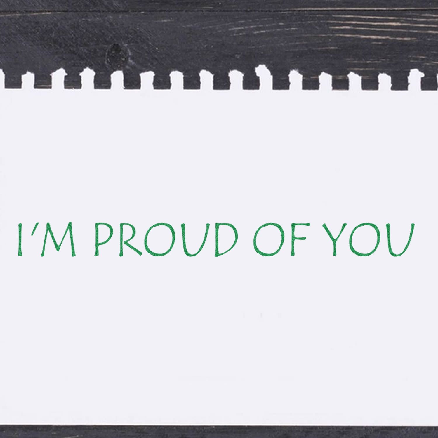 A white paper stamped with green ink using the I'm Proud Of You Rubber Stamp on a dark wooden surface.