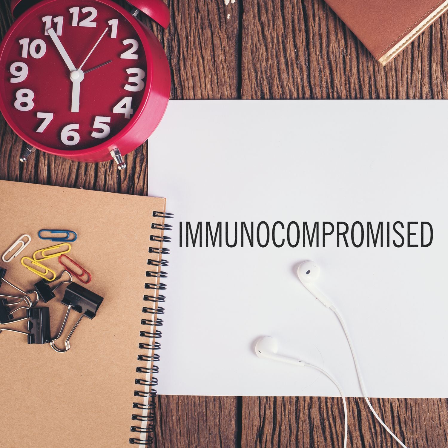 Large Immunocompromised Rubber Stamp on a desk with a red clock, notebook, paper clips, binder clips, and earphones.