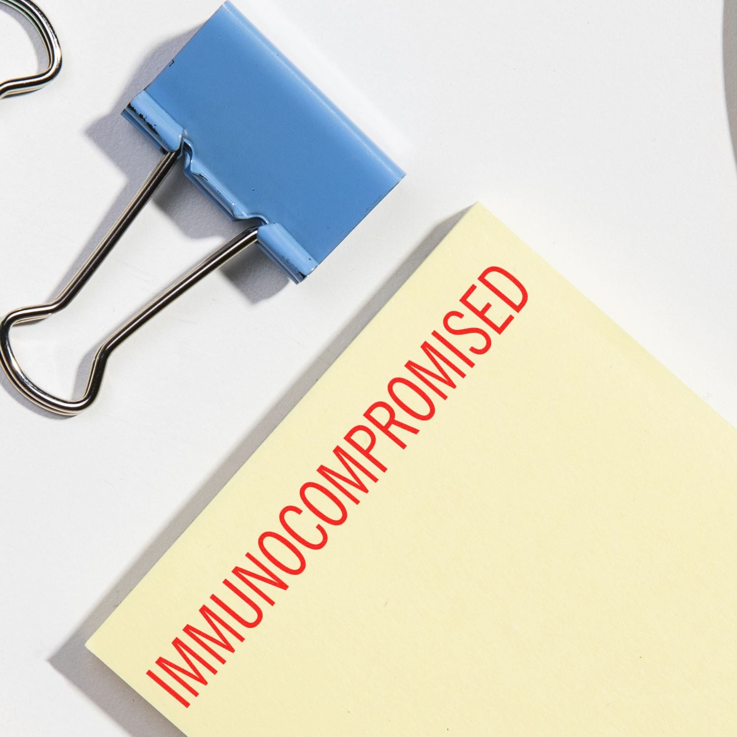 Immunocompromised Rubber Stamp in red ink on a yellow paper, next to a blue binder clip on a white background.