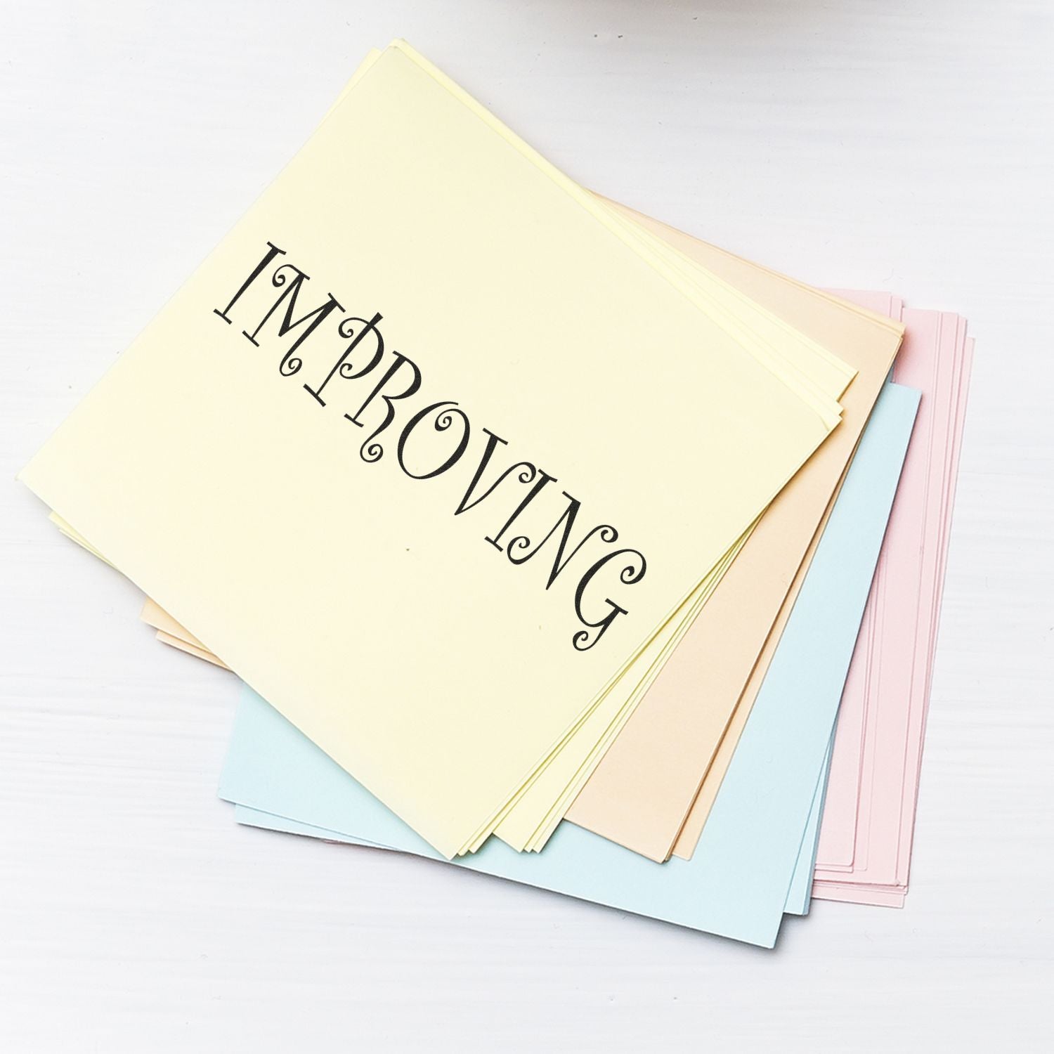 A stack of pastel-colored papers stamped with IMPROVING using the Improving Rubber Stamp, placed on a white surface.