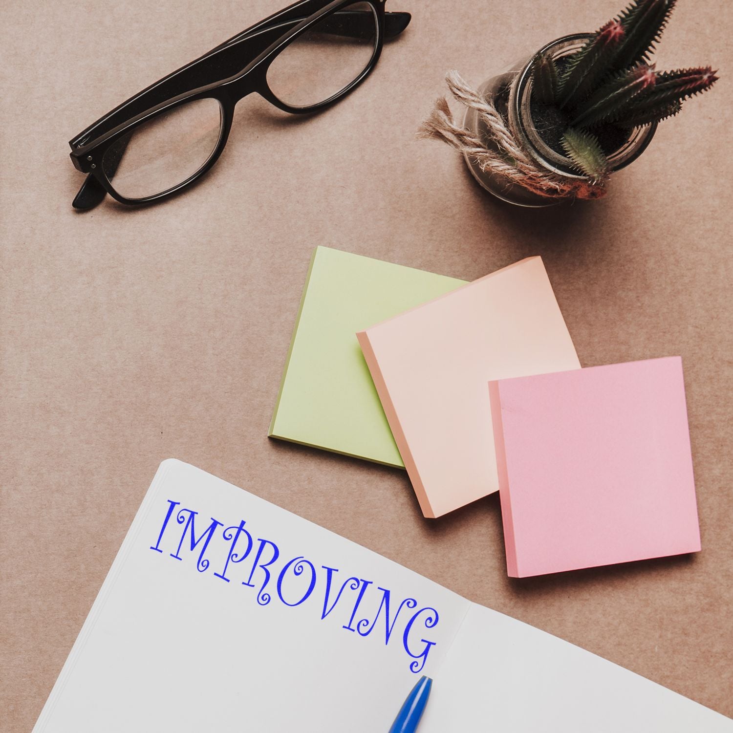 Large Pre-Inked Improving Stamp in use on a notebook, surrounded by colorful sticky notes, glasses, and a small potted plant.