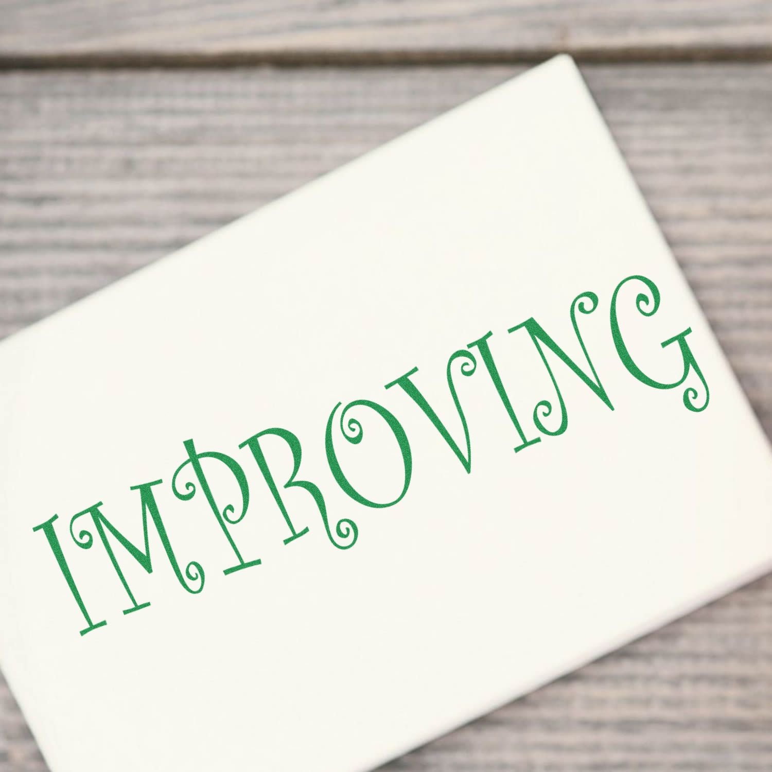 Improving Rubber Stamp in use, displaying the word IMPROVING in green ink on a white surface, placed on a wooden background.