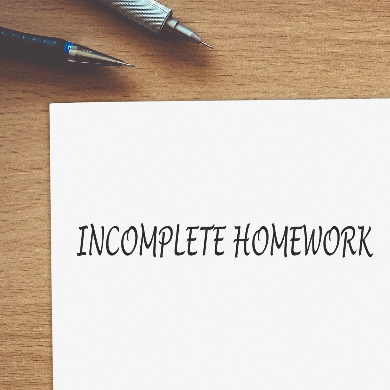 Large Pre-Inked Incomplete Homework Stamp on a white paper with INCOMPLETE HOMEWORK text, placed on a wooden desk with two pens.